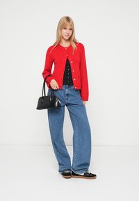 Red knitted cardigan with gold buttons, black top, loose blue denim jeans, and black handbag. Model wears black and tan sneakers.