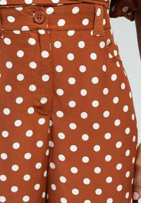 Tissu marron avec de grands pois blancs, doté d'un bouton frontal et de poches, mettant en valeur une texture lisse et un design ajusté.