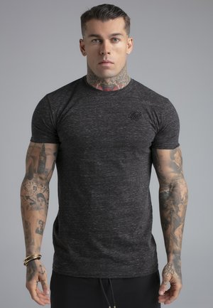 Homme musclé avec des tatouages sur le cou et les bras, debout face à la caméra, portant un t-shirt gris foncé à manches courtes ajusté sur un fond gris uni.