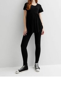 Žena sedící v černé tunice a legínách stojí proti světlému šedému zdi, oblečená v černých vysokých teniskách.