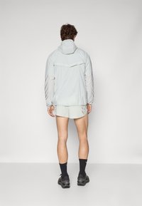 Chaqueta zip-up gris claro con capucha, que presenta rayas oscuras contrastantes en las mangas; combinada con pantalones cortos a juego y calcetines negros. De pie desde atrás.