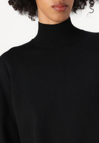 Svart genser med høy hals og ribbestruktur, med en løs silhuett, laget av mykt strikkemateriale.