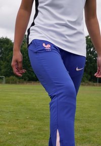 Mėlynos sportinės kelnės su rožiniu prancūzišku gaidžio logotipu ir balta Nike swoosh. Yra aptempto silueto dizainas ir šoninės skylutės šalia kulkšnų.