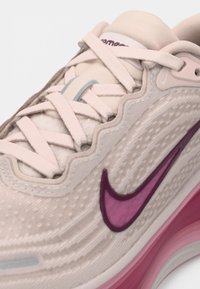 Beige sportcipő texturált felsőrésszel, rózsaszín színátmenetes talppal és kiemelkedő lila Swoosh logóval. Jellemzője a lapos, fűzős kialakítás sima díszítésekkel.