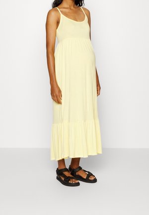 Kobieta w ciąży w jasnopomarańczowej, bezrękawowej sukience maxi z falbaną na dole i czarnych sandałach z odkrytymi palcami, stojąca na jednolitym tle.