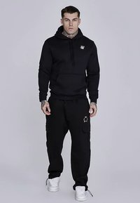 Felpa con cappuccio nera con tasca anteriore, indossata con pantaloni cargo neri abbinati. Presenta un piccolo logo sulla felpa e un accento dorato sui pantaloni.