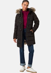 Svart vadderad jacka med pälsfodrad huva, tryckknappar och dragkedja, sidofickor och knälång design, kombinerad med en vinröd topp och jeans.