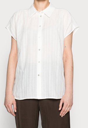 Osoba ubrana w białą koszulę z krótkim rękawem zapinaną na guziki z subtelnymi pionowymi paskami, w połączeniu z ciemnobrązowymi spodniami, stojąca na jasnym tle.