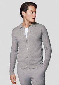 Jasnoszary kardigan z kołnierzem, z przodu zapinany na guziki, z prążkowanymi mankietami i dołem. Noszony na białej koszuli o gładkiej fakturze.