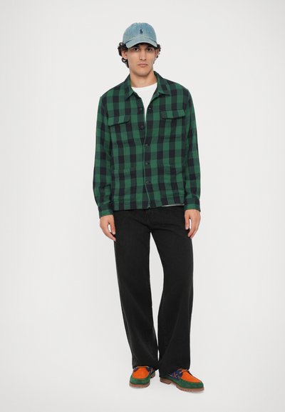 Camisa em padrão xadrez verde e preto com dois bolsos no peito, combinada com calças pretas e sapatos texturizados multicoloridos. Boné de ganga com detalhe do logótipo.