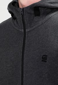Sweat-shirt zippé gris foncé en tissu texturé. Il présente un col montant, une fermeture éclair noire et des détails de logo subtils. Coupe lisse et athlétique.