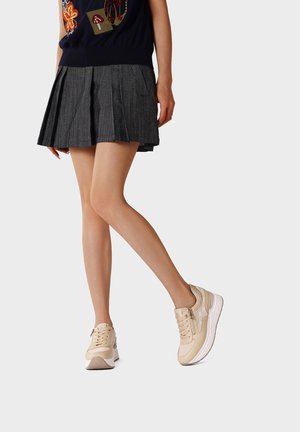 Donna con sneakers beige con zeppa e gonna grigia plissettata, in piedi con le gambe incrociate su uno sfondo chiaro.