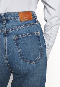 Vysoce pasové modré džíny z denimu s koženou nášivkou na pásu. Mezi další prvky patří zadní kapsy a viditelné detaily prošívání.