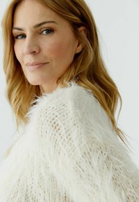 Vrouw met lichtbruin haar, gekleed in een wit, zacht, gebreid truitje, die zachtjes in de camera kijkt tegen een effen lichte achtergrond.