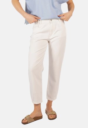 Kvinne iført off-white høytlivs rette bukser med beige sandaler med spenne, justerer lys blå skjorte stukket inn i buksene.