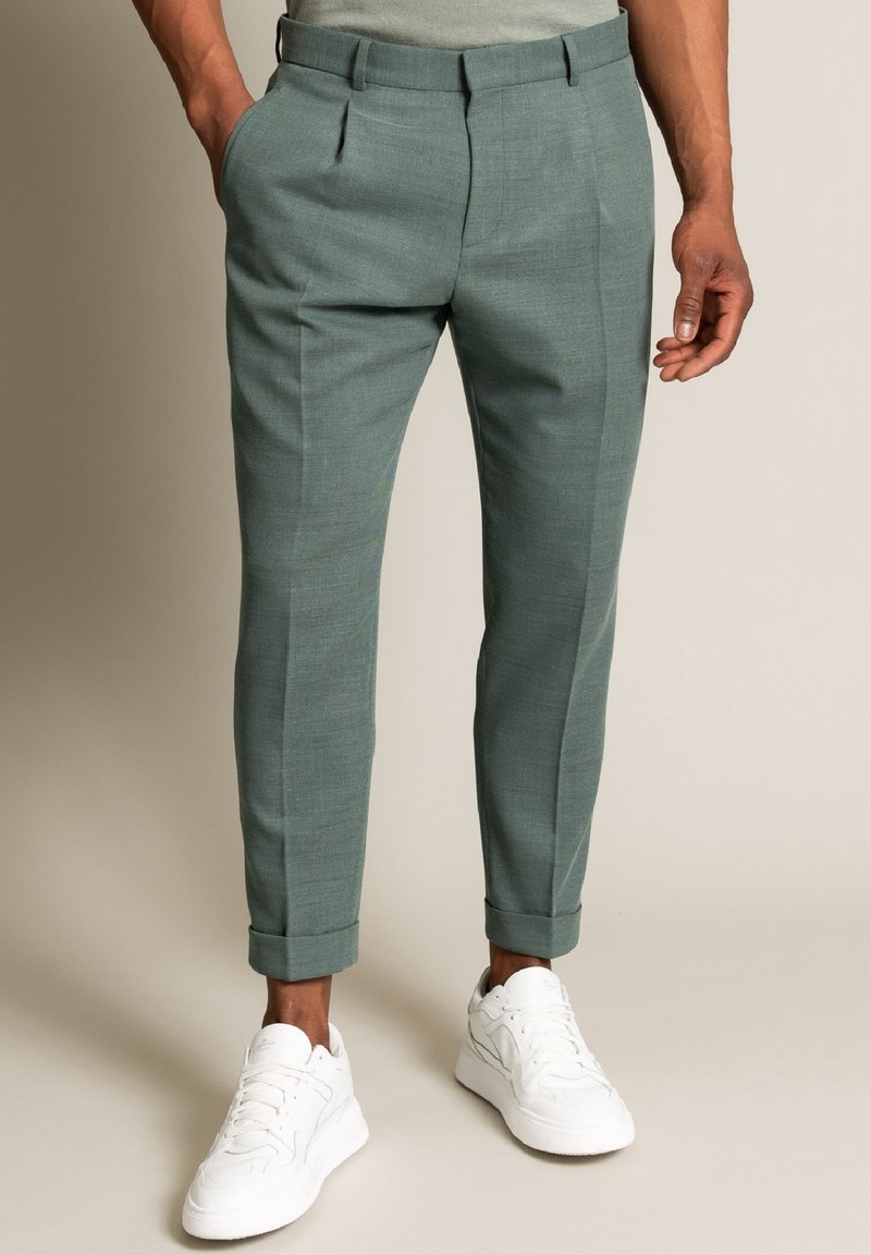 Hellgrüne, maßgeschneiderte Hosen mit einer strukturierten Oberfläche, versehen mit vorderen Bügelfalten und aufgekrempelten Bündchen, kombiniert mit weißen Turnschuhen.