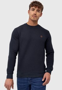 Navy textured sweater with a round neckline, long sleeves, and ribbed cuffs. Features a small brown logo on the chest.