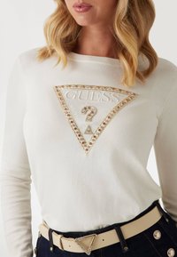 Camiseta blanca de manga larga con un logo "GUESS" acentuado con strass en forma de triángulo. Combinada con un cinturón beige con una hebilla triangular sobre un denim oscuro.