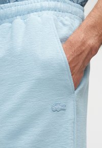 Shorts bleu clair en tissu texturé, avec un logo brodé sur le côté gauche et une poche latérale. Ceinture élastique visible.