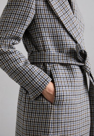 Houndstooth-patterned coat in beige and blue tones, featuring a belted waist, wide lapels, and side pockets. Textured fabric close-up.