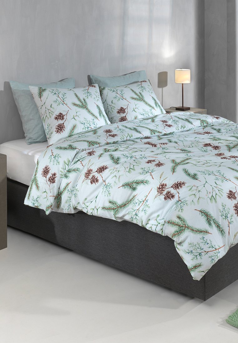 Tweepersoonsbed met een grijze basis en beddengoed met botanische print met dennenappels en groene bladeren, naast een nachtkastje met een brandende lamp in een minimale kamer.