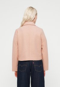 Κοντό ροζ μάλλινο σακάκι με γιακά με εγκοπή. Λείες υφές και ελάχιστα ραφές, σε συνδυασμό με ψηλόμεσα σκούρα τζιν παντελόνια με πίσω τσέπες.