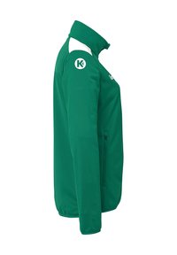 Grøn jakke med lynlåslukning, høj krave og hvide accenter. Har et rundt logo på skulderen og en sidelomme med lynlås. Glat tekstur.