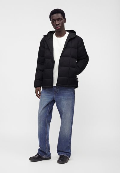 Jeune homme portant une doudoune noire, une chemise blanche, un jean bleu large et des chaussures noires, debout devant un fond uni.
