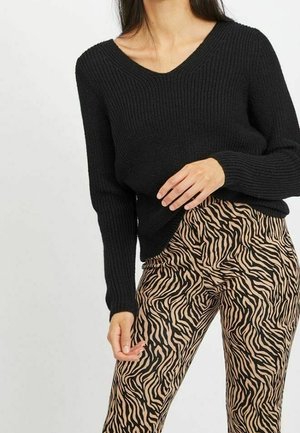 Femme portant un pull à col en V côtelé noir et un pantalon beige à rayures zébrées noires, posant avec une main près de la taille.