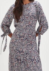 Granatowa sukienka w wzór paisley z misternymi białymi i czerwonymi wzorami, z rękawami o długości trzech czwartych z kokardkami oraz luźnym krojem.