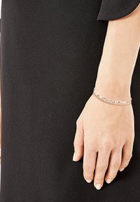 Pulsera de plata con múltiples hilos, decorada con pequeñas cuentas y un charm de símbolo de infinito, llevada en una mano de piel clara, sobre un fondo negro.