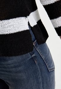 Pull en maille rayé noir et blanc avec fente latérale, associé à un jean en denim bleu dévoilant les détails visibles des poches et des coutures.