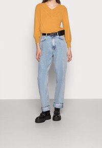 Mosterdgele gebreide trui met een V-hals, gecombineerd met lichtblauwe high-waisted jeans die opgerold zijn bij de manchetten en zwarte platform enkellaarsjes.