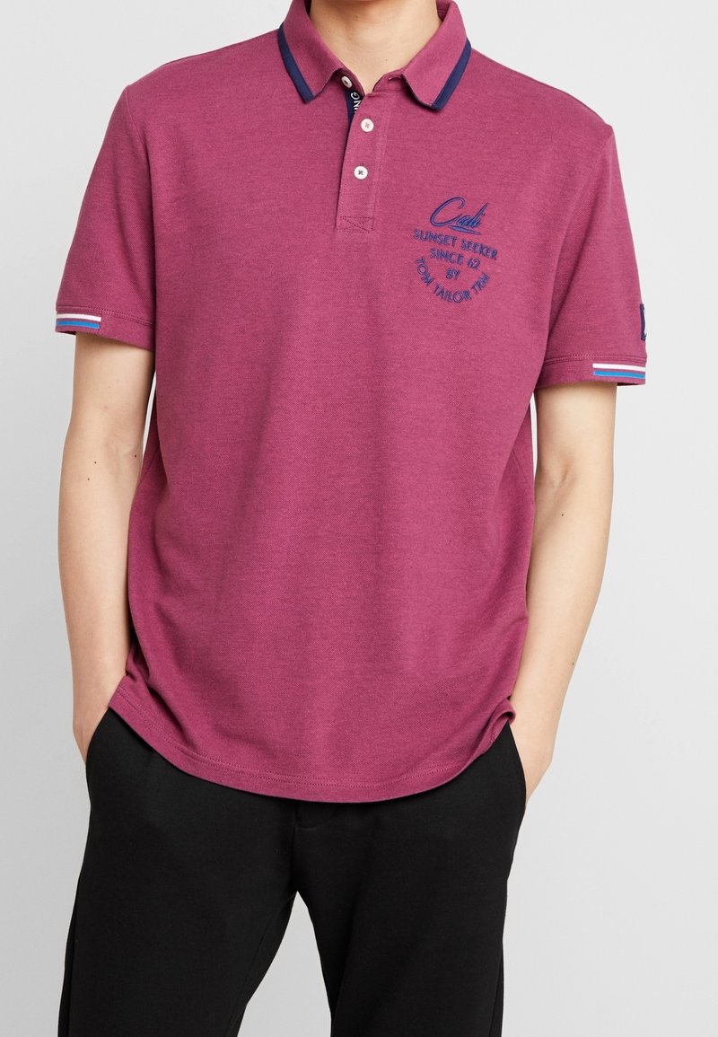 Bordeauxfärgad polo skjorta med krage, två knappar, blå detaljer på kragen och ärmarna, samt en broderad logotyp på bröstet.