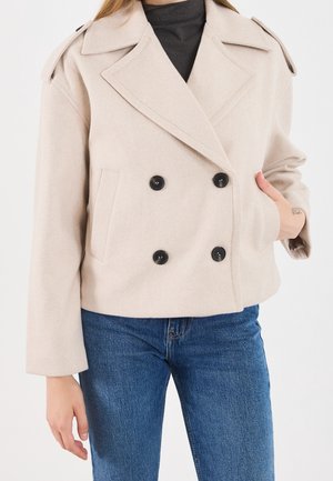 Femme portant une veste en laine beige à double boutonnage avec des boutons noirs, un col roulé noir et un jean bleu, main dans la poche de la veste.