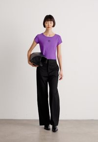 Purple short-sleeve t-shirt with a logo, paired with high-waisted black wide-leg trousers and a black handbag, highlighting a modern outfit.