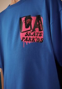 Niebieska bawełniana koszulka z dużym nadrukiem graficznym w czerni i różu, z tekstem „LA SKATE PARK’95”, o luźnym kroju i okrągłym dekolcie.