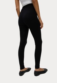 Sorte leggings lavet af strækbart materiale, med en høj talje og en tætsiddende pasform, parret med flade sorte sko.