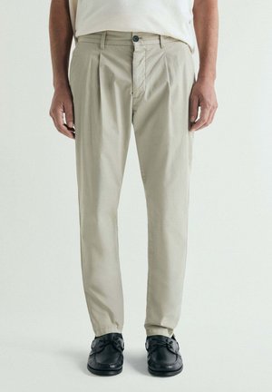 Hombre con pantalones plisados beige y zapatos de cuero negros, de pie frente a un fondo liso.