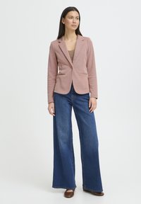 Blazer rosa texturizado con cierre de un solo botón, combinado con vaqueros de mezclilla azul de pierna ancha. Modelo posando frente a un fondo blanco liso.