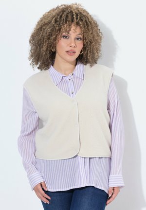 Kvinde med krøllet hår iført en cremefarvet vest over en lyse-lilla stribet skjorte og blå jeans mod en ensfarvet hvid baggrund.