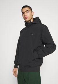 Veste à capuche légère noire avec des poignets et une ceinture élastiques. Dotée d'une fermeture éclair à l'avant et d'un logo sur la poitrine. Texture lisse.