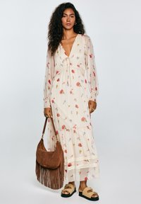 Maxi-jurk in lichtbeige, met bloemmotieven in roze en blauw, diepe V-hals, strik aan de voorkant, lange mouwen en kantaccenten. Gekocht met een bruine franjerugzak.