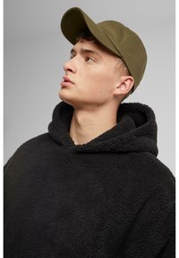 Hoodie en polaire noir avec une finition texturée et une grande capuche, associé à une casquette de baseball verte olive. Le mannequin regarde vers le haut.