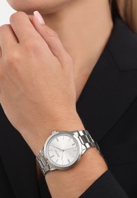 Srebrny zegarek metalowy z okrągłą tarczą i gładką białą cyferblatą. Posiada kryształowe akcenty wokół obręczy oraz wypolerowaną bransoletę.