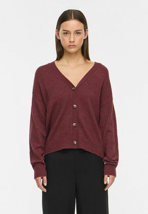 Jeune femme aux cheveux bruns raides porte un cardigan bordeaux boutonné et un pantalon noir, debout devant un fond blanc uni.