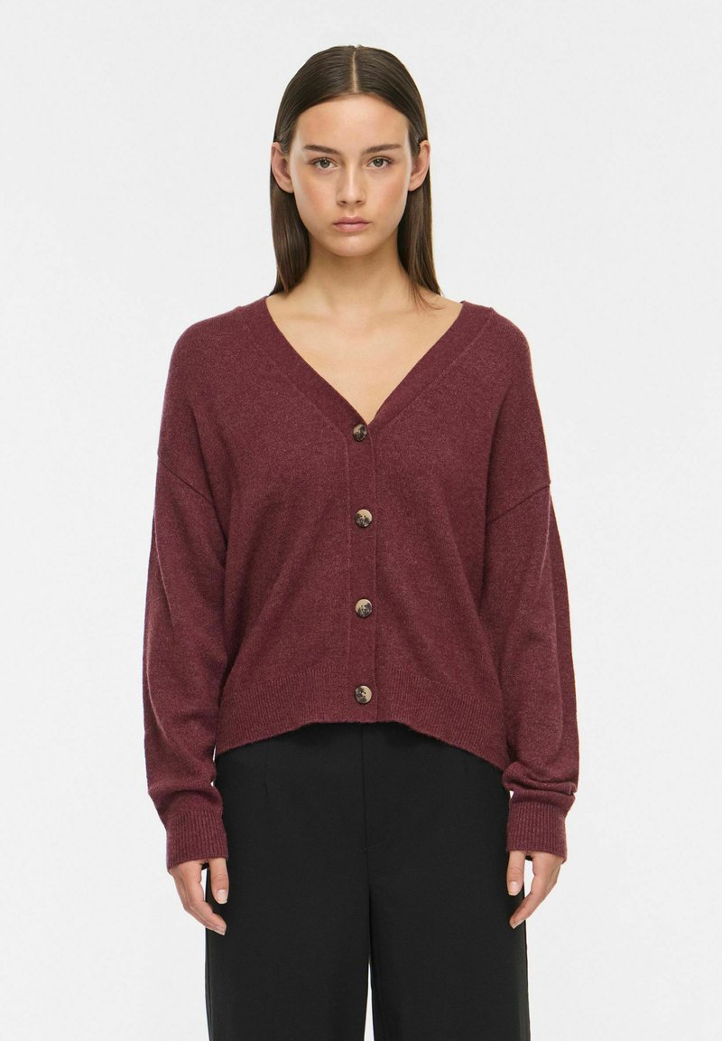 Jeune femme aux cheveux bruns raides porte un cardigan bordeaux boutonné et un pantalon noir, debout devant un fond blanc uni.