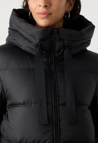 Zwarte pufferjas met hoge kraag, ritssluiting en dikke padding. Het heeft twee lange zwarte stoffen banden met logo-accenten.
