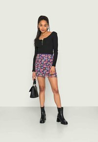 Blusa preta de mangas longas com detalhe de fecho, combinada com uma saia mini de padrão floral e botas pretas até ao tornozelo. Modelo segura uma pequena mala preta.