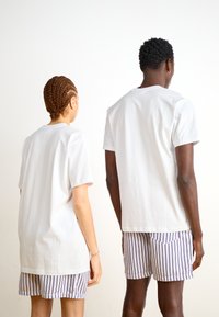 Hvide bomulds t-shirts med korte ærmer, båret med stribede shorts, set fra bagfra. Simpelt design, lige snit, ren finish.